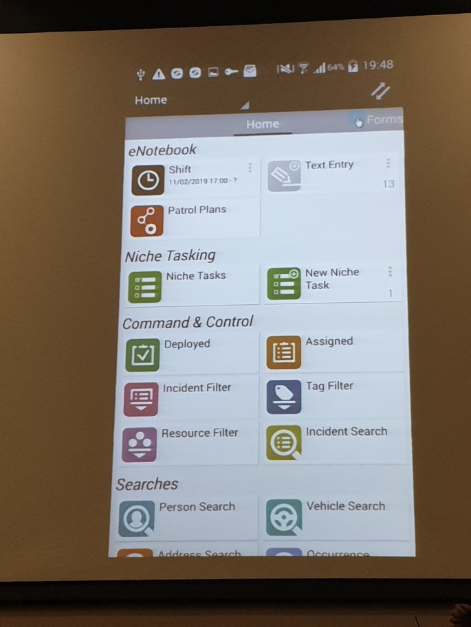 An image on an overhead projector of the police mobile app, with sections for eNotebook, Niche Tasking, Command and Control and Searches. The latter contains the fingerprint scanning app, which they call Person Search.